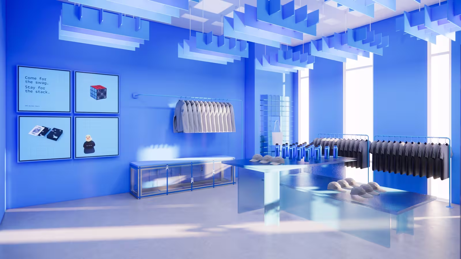 3D rendering of the AWS Builder Loft swag shop, bathed entirely in blue. The room features clothing racks with grey hoodies and black t-shirts, display tables with baseball caps and blue water bottles, a tote bag on a wall hook, and a glass display case. A four-panel wall display reads "Come for the swag. Stay for the stack." alongside images of merchandise and a pixel-art Rubik's cube. Sculptural geometric cube installations hang from the ceiling in translucent blue, echoing the Builder Loft design language. Floor-to-ceiling windows let in soft light, and the monochromatic blue palette gives the space a bold, immersive retail feel.
